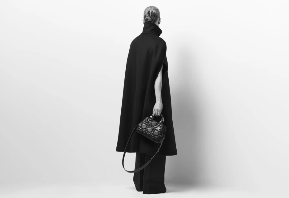 A woman wearing a black cape-like coat and wide-leg pants stands against a white backdrop holding a small, ornate black purse.