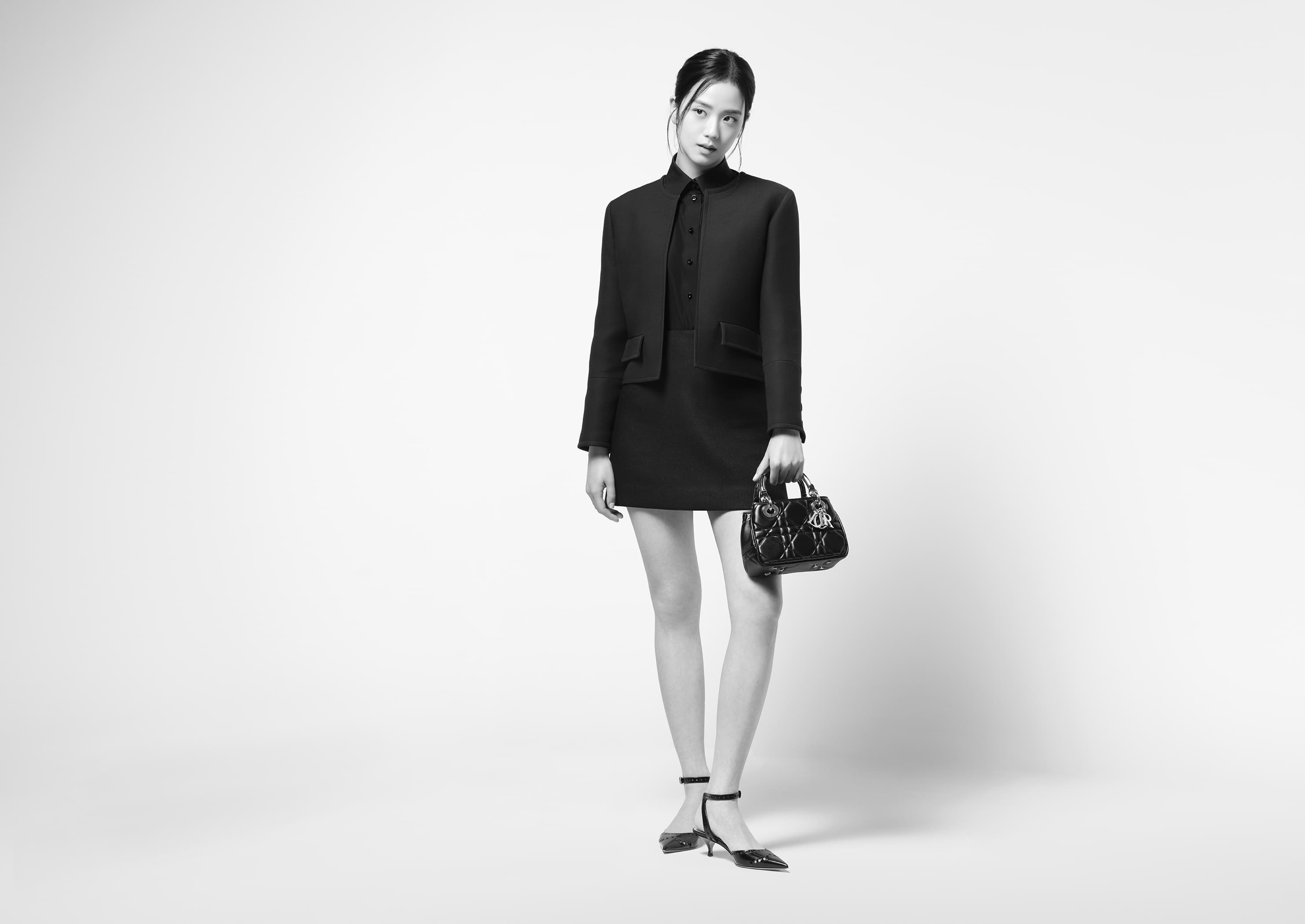A full-length black and white studio shot presents a stylish Asian woman in a dark skirt suit, complemented by a matching purse and heels.
