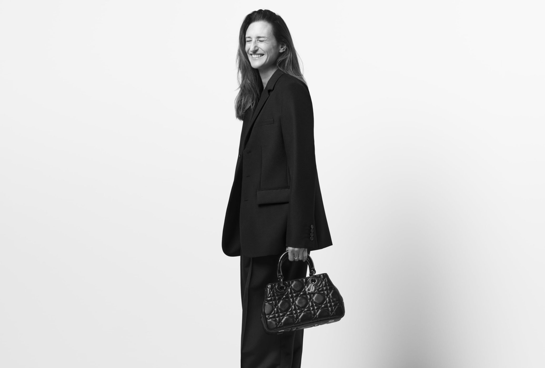 Rachele Regini smiling in a black tailored suit and holding a black quilted handbag against a white studio background.