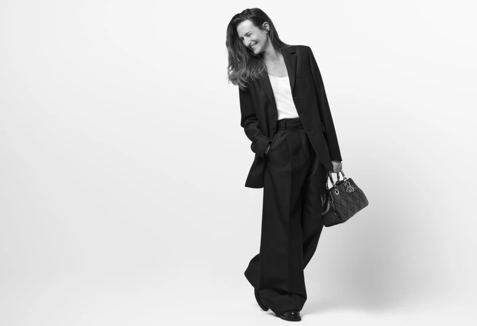 Monochrome studio shot of a woman in a dark suit and wide-leg pants, holding a quilted handbag and smiling in a relaxed pose.