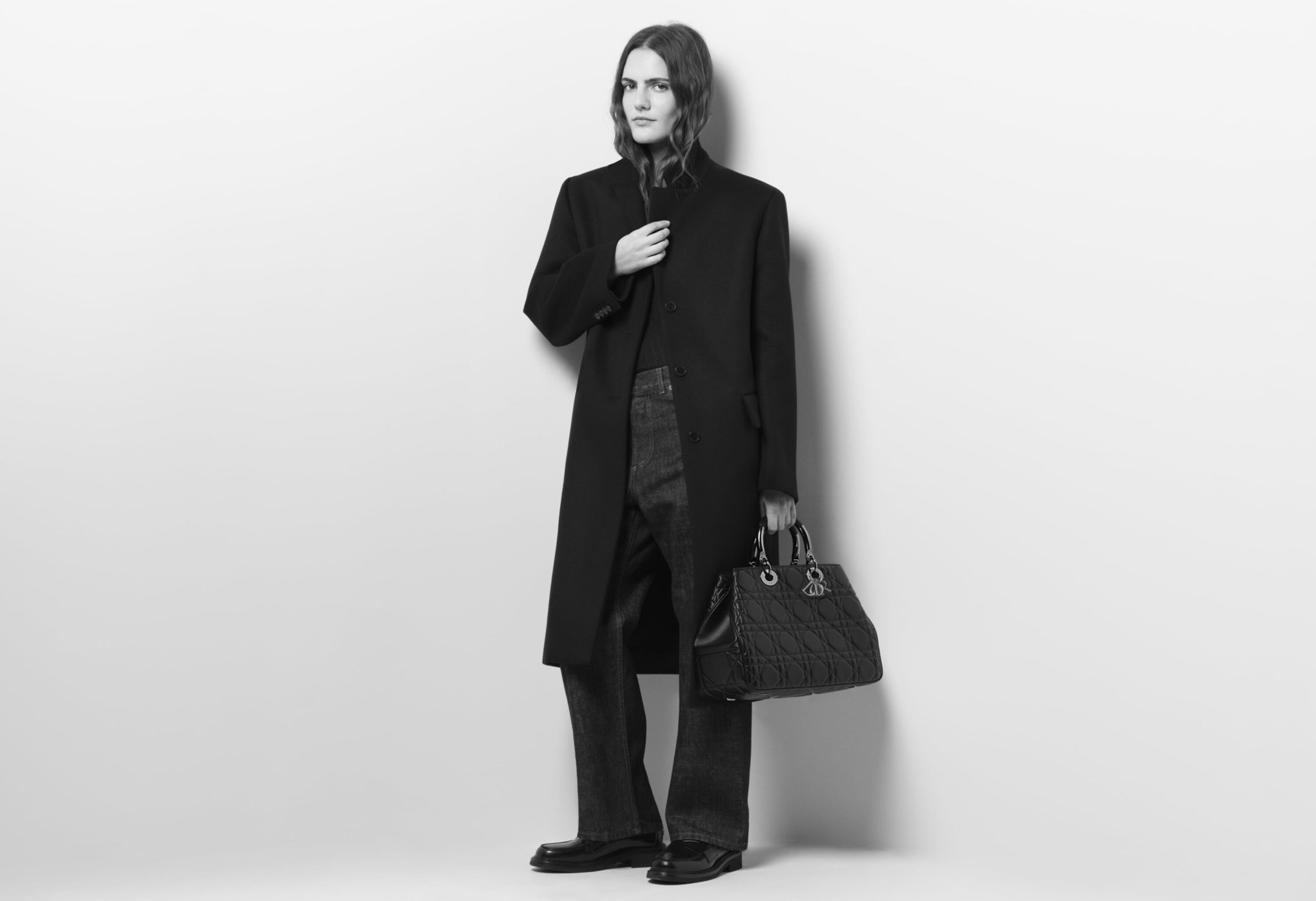 Black and white full-shot of a person in a black coat and jeans, holding a Dior handbag against a light wall.