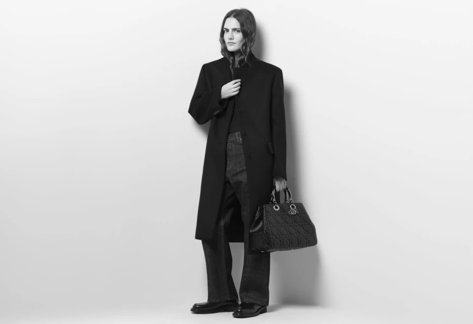 Black and white full-shot of a person in a black coat and jeans, holding a Dior handbag against a light wall.