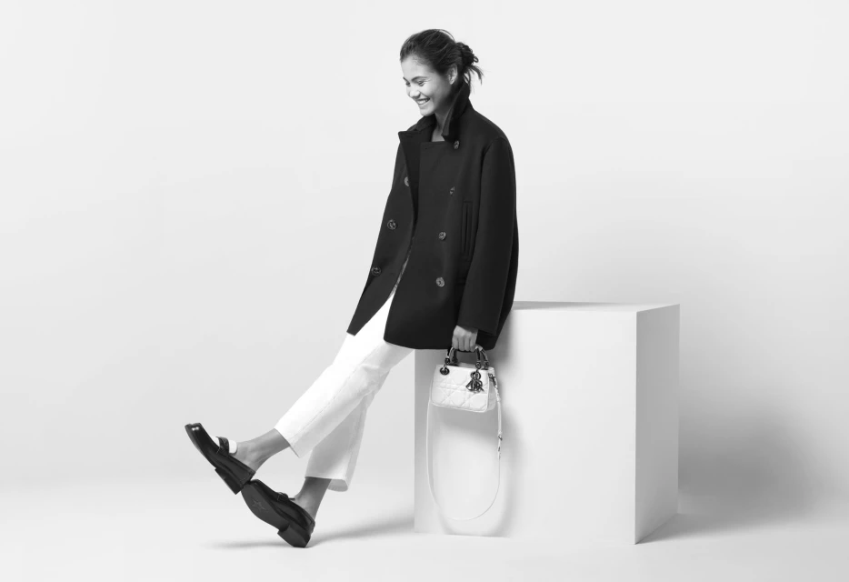 A stylish adult woman in a black coat and white pants sits on a white cube, holding a white purse against a plain white background.