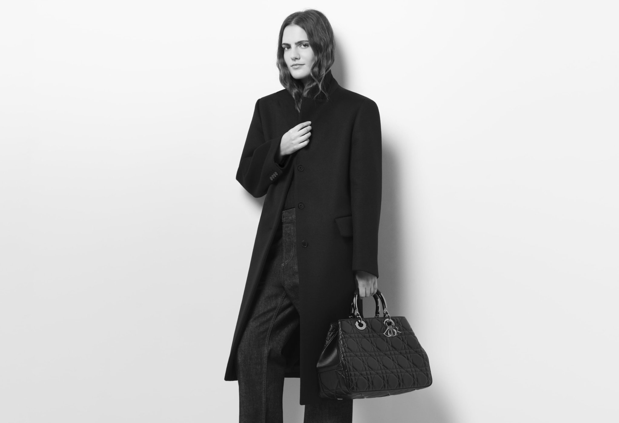 Elegant woman in a black coat holds a quilted black handbag against a white background in a studio, creating a high-fashion aesthetic.