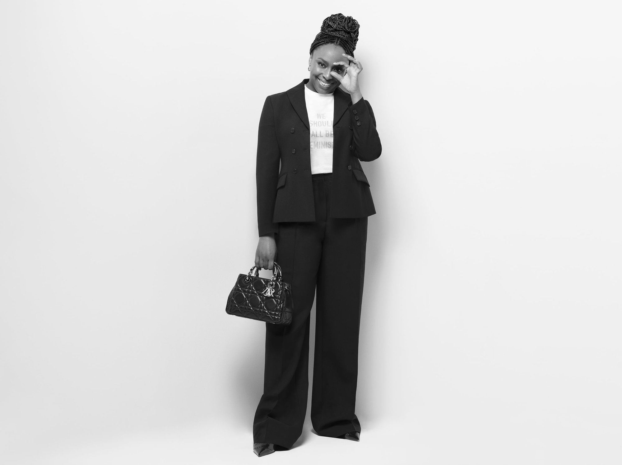 Full shot of an adult woman smiling and posing indoors wearing a black suit, a white t-shirt, holding a black purse, in a photo studio.