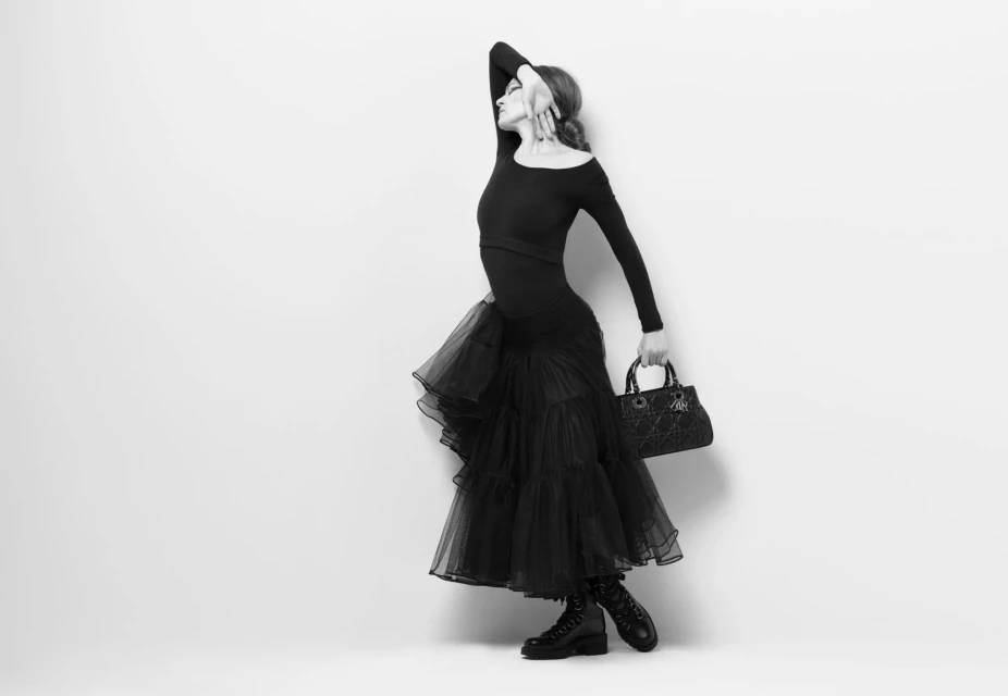 Fashion model in a black dress, boots, and holding a handbag poses in a studio with a white backdrop, side view in black and white.