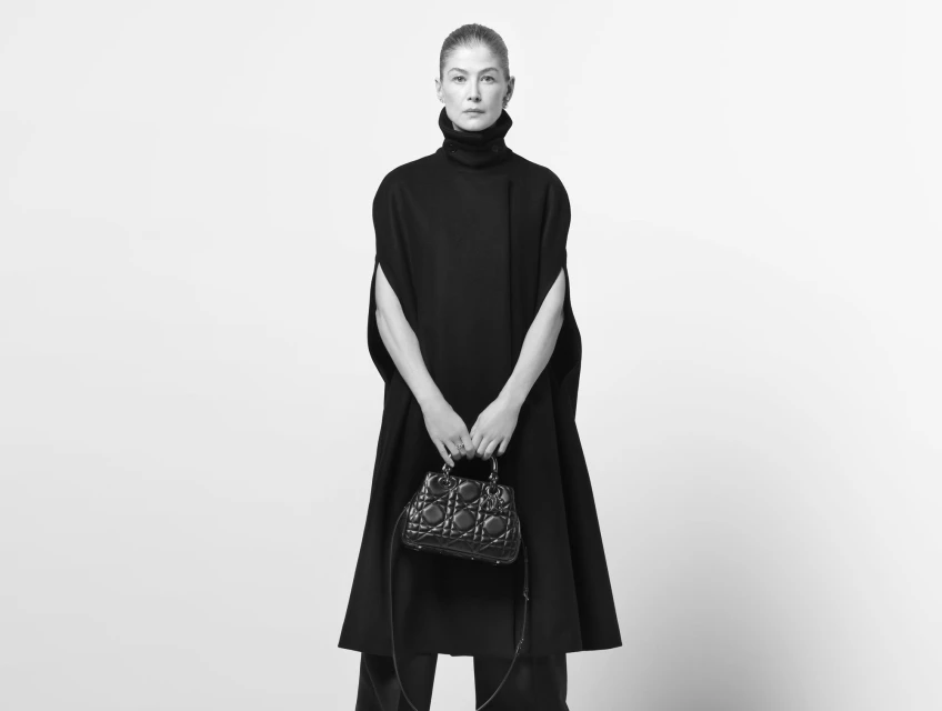 Elegant woman in a black cape dress poses in a studio with a structured leather handbag, conveying classic fashion and style.