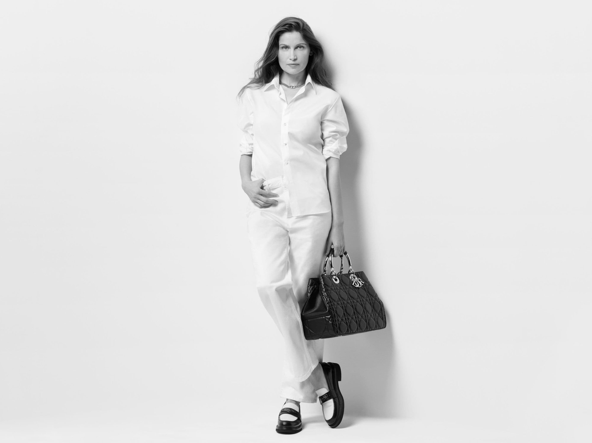 Elegant woman in white shirt and pants poses with a black leather Lady Dior handbag in a high-key fashion shot against a white wall.