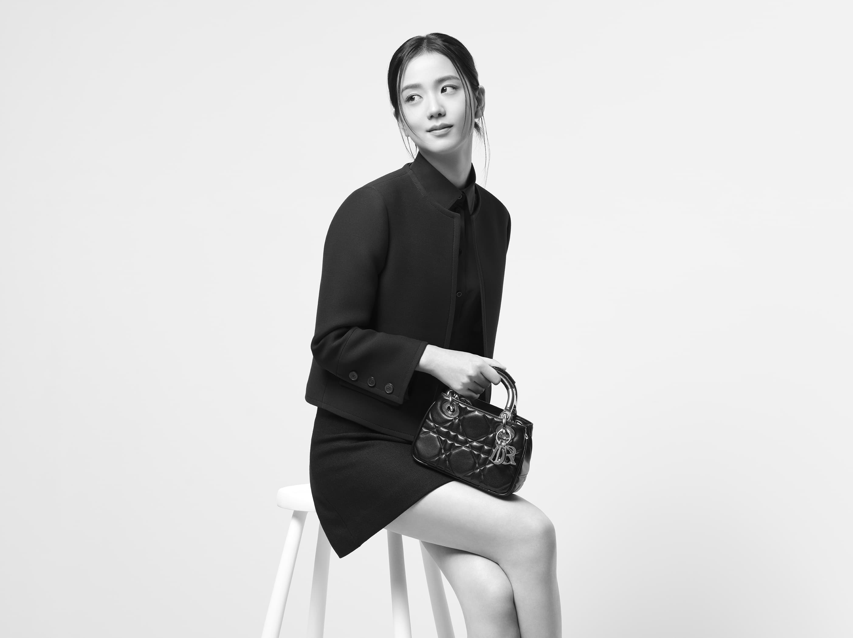 Monochrome portrait of a seated young woman with dark hair in a black jacket and skirt holding a Lady Dior handbag, looking off-camera.