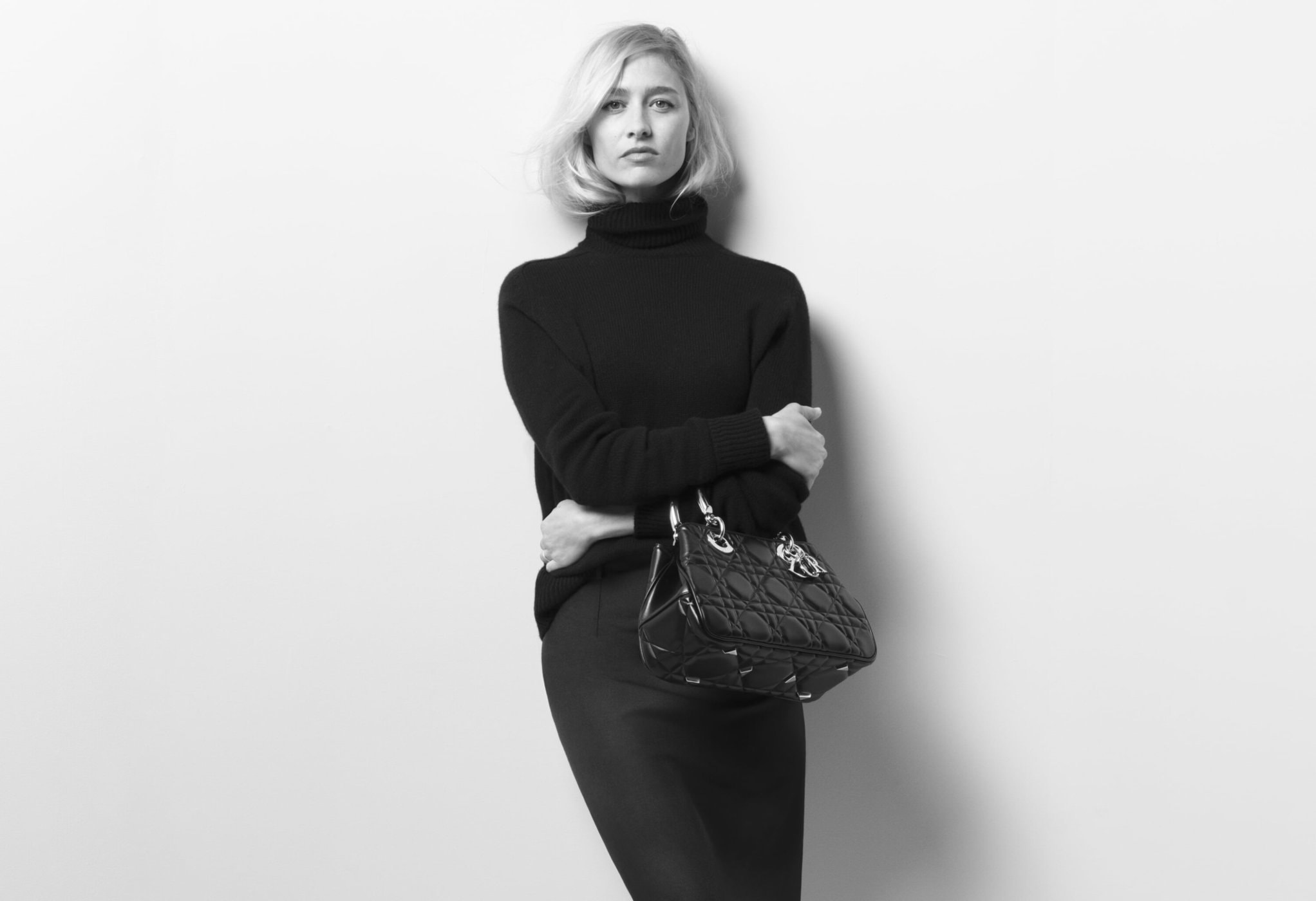 Black and white studio shot of a blonde woman in a black turtleneck and skirt, holding a purse, against a white backdrop.