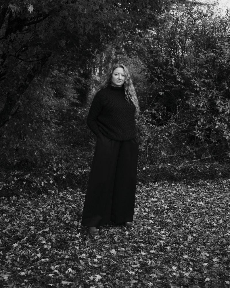 Full-length portrait of a young woman with long curly hair in a black sweater and wide-leg pants standing in a leafy forest. 