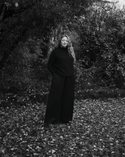 Full-length portrait of a young woman with long curly hair in a black sweater and wide-leg pants standing in a leafy forest. 