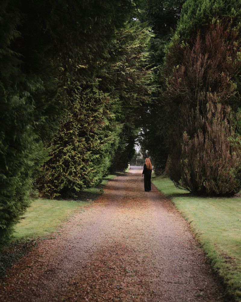 Woman walking along a gravel path lined with tall, green trees in a park. The natural setting and solitary figure create a tranquil scene.
