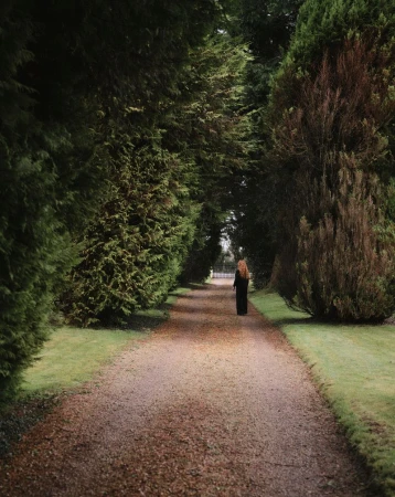 Woman walking along a gravel path lined with tall, green trees in a park. The natural setting and solitary figure create a tranquil scene.