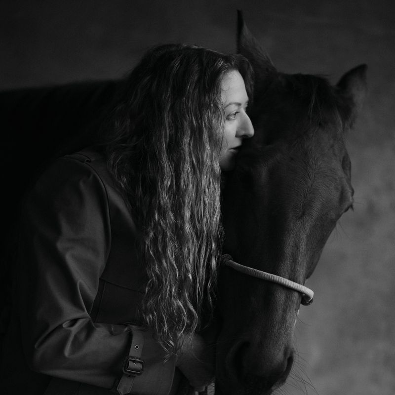 Monochrome portrait of a woman with long curly hair standing next to a horse with a dark coat in an indoor stable setting.