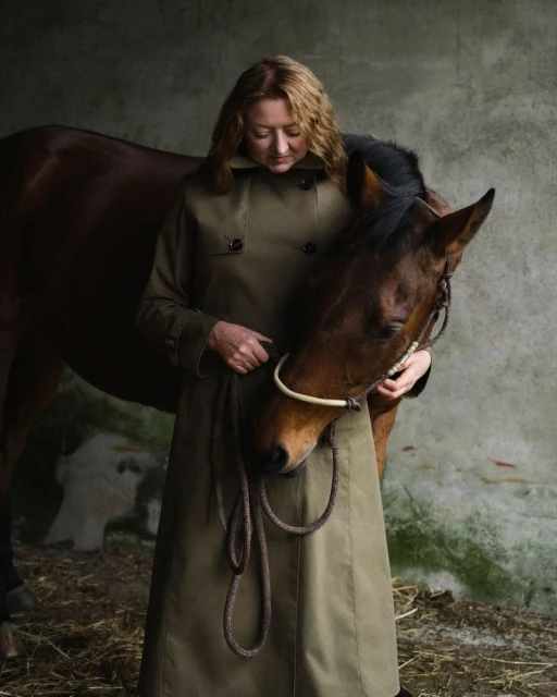 Woman in trench coat with horse in indoor stable. Portrait shot features brown horse and adult woman looking down. Muted color palette.