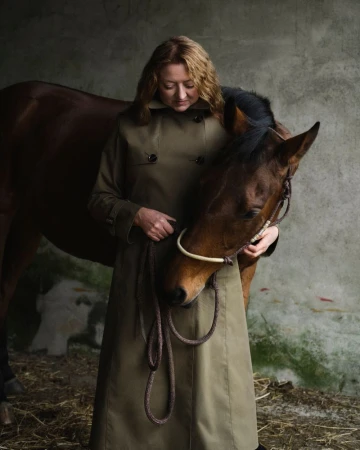 Woman in trench coat with horse in indoor stable. Portrait shot features brown horse and adult woman looking down. Muted color palette.