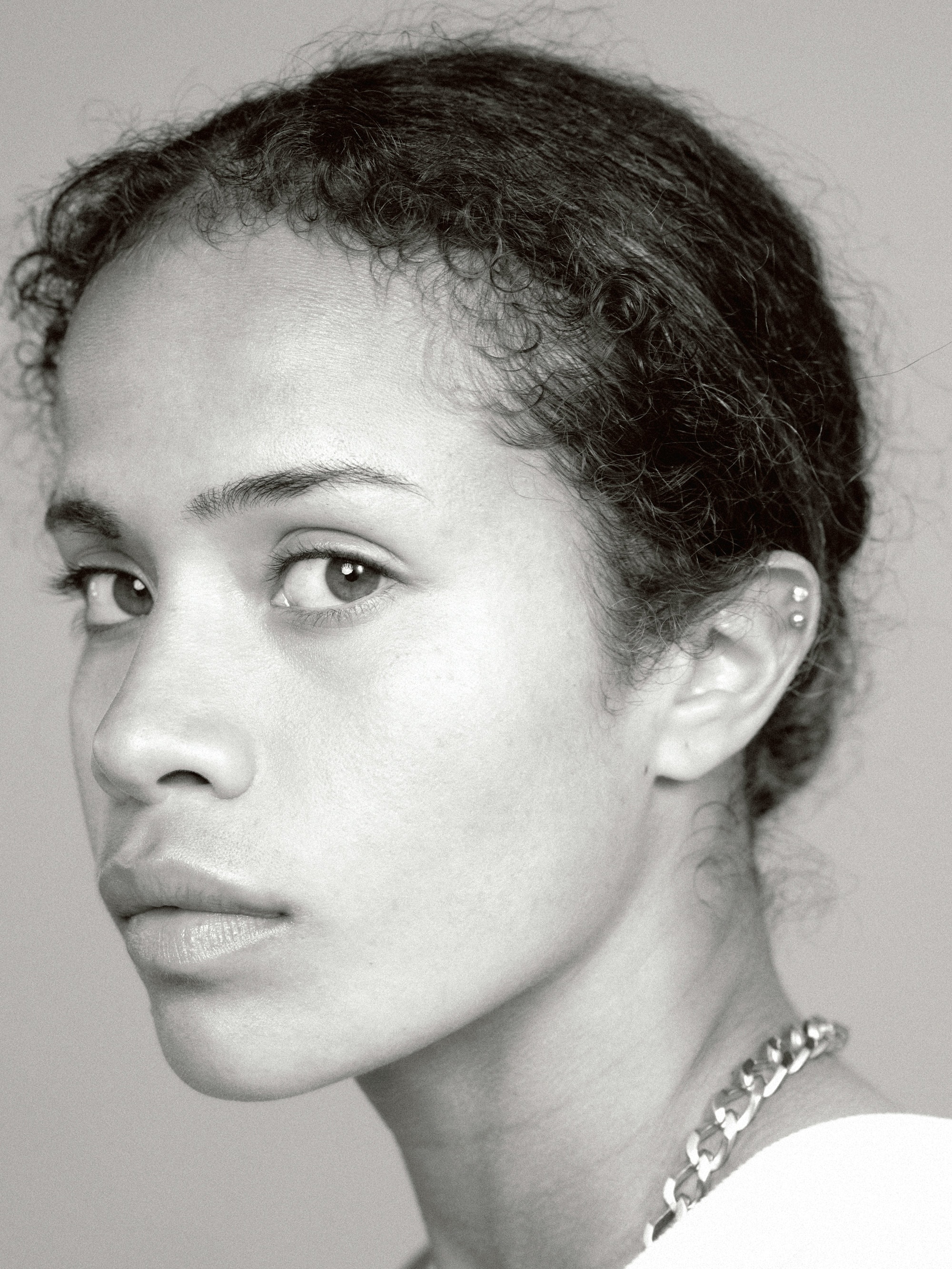 Close-up portrait of a young adult woman with curly hair, wearing a chain necklace, in a black and white studio setting.