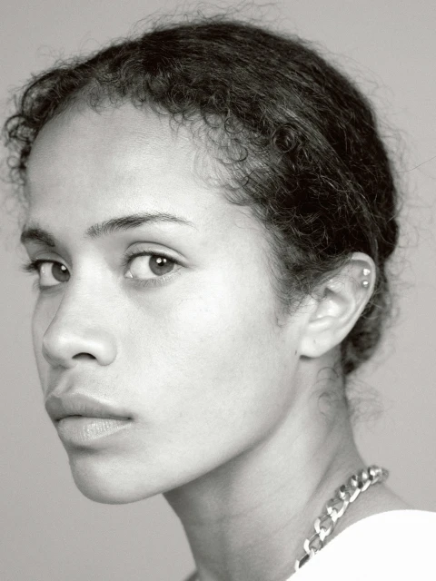 Close-up portrait of a young adult woman with curly hair, wearing a chain necklace, in a black and white studio setting.