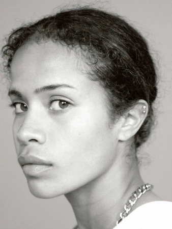 Close-up portrait of a young adult woman with curly hair, wearing a chain necklace, in a black and white studio setting.