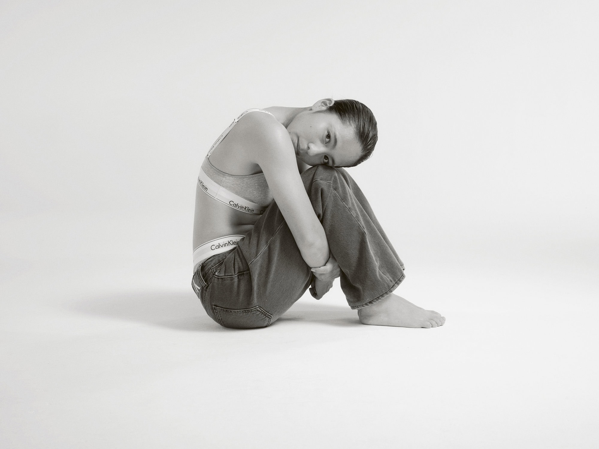 Woman in Calvin Klein bra and jeans sitting in a photo studio with legs up, hugging them, looking at the camera.