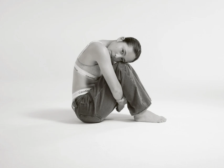 Woman in Calvin Klein bra and jeans sitting in a photo studio with legs up, hugging them, looking at the camera.