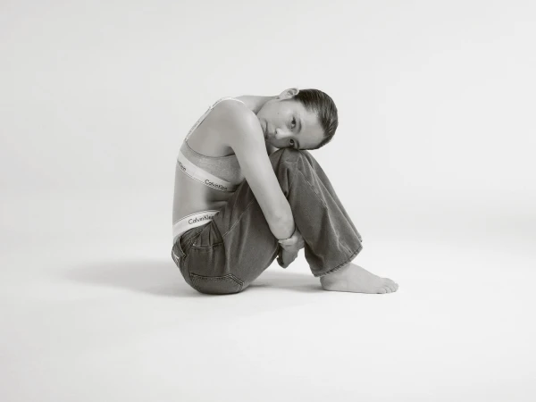 Woman in Calvin Klein bra and jeans sitting in a photo studio with legs up, hugging them, looking at the camera.