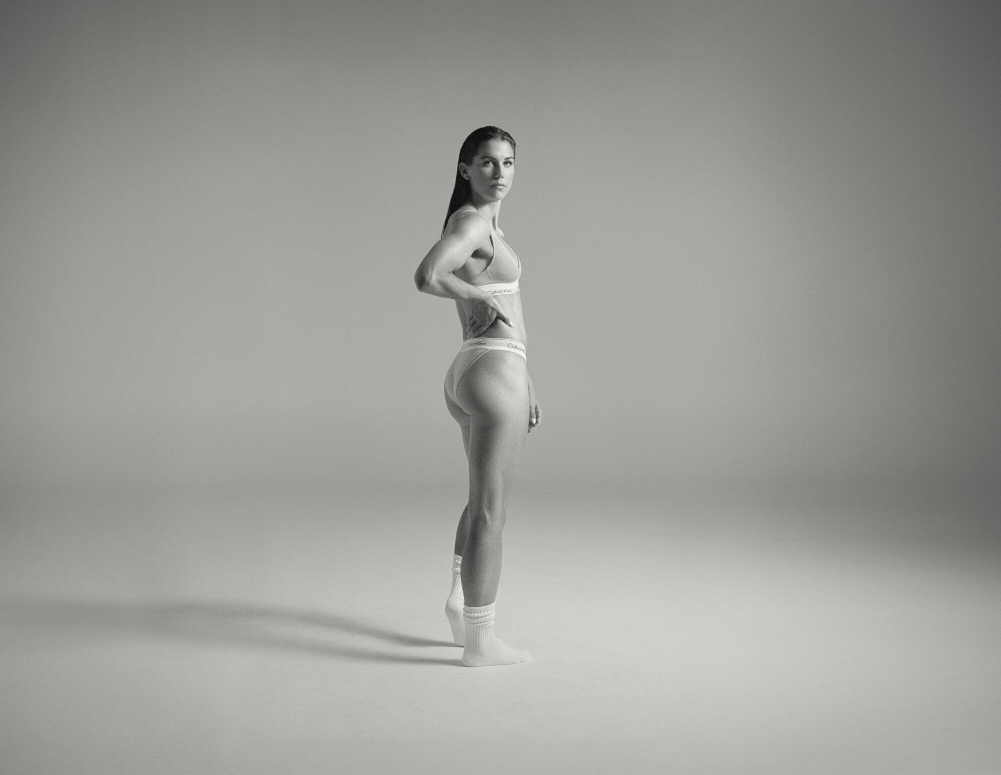 Side view of a slender woman in underwear and socks standing in a brightly lit studio, posing with her hand on her hip.