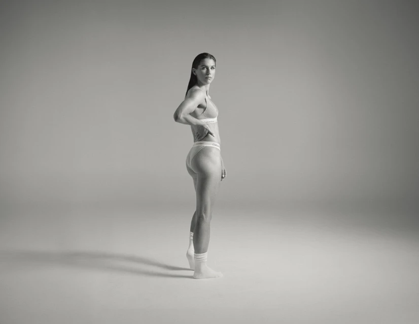 Side view of a slender woman in underwear and socks standing in a brightly lit studio, posing with her hand on her hip.