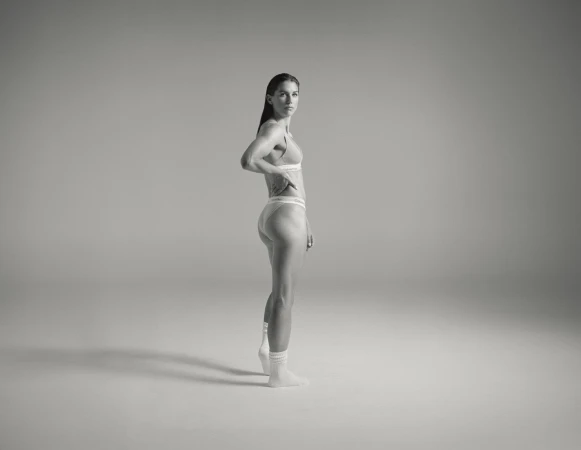 Side view of a slender woman in underwear and socks standing in a brightly lit studio, posing with her hand on her hip.