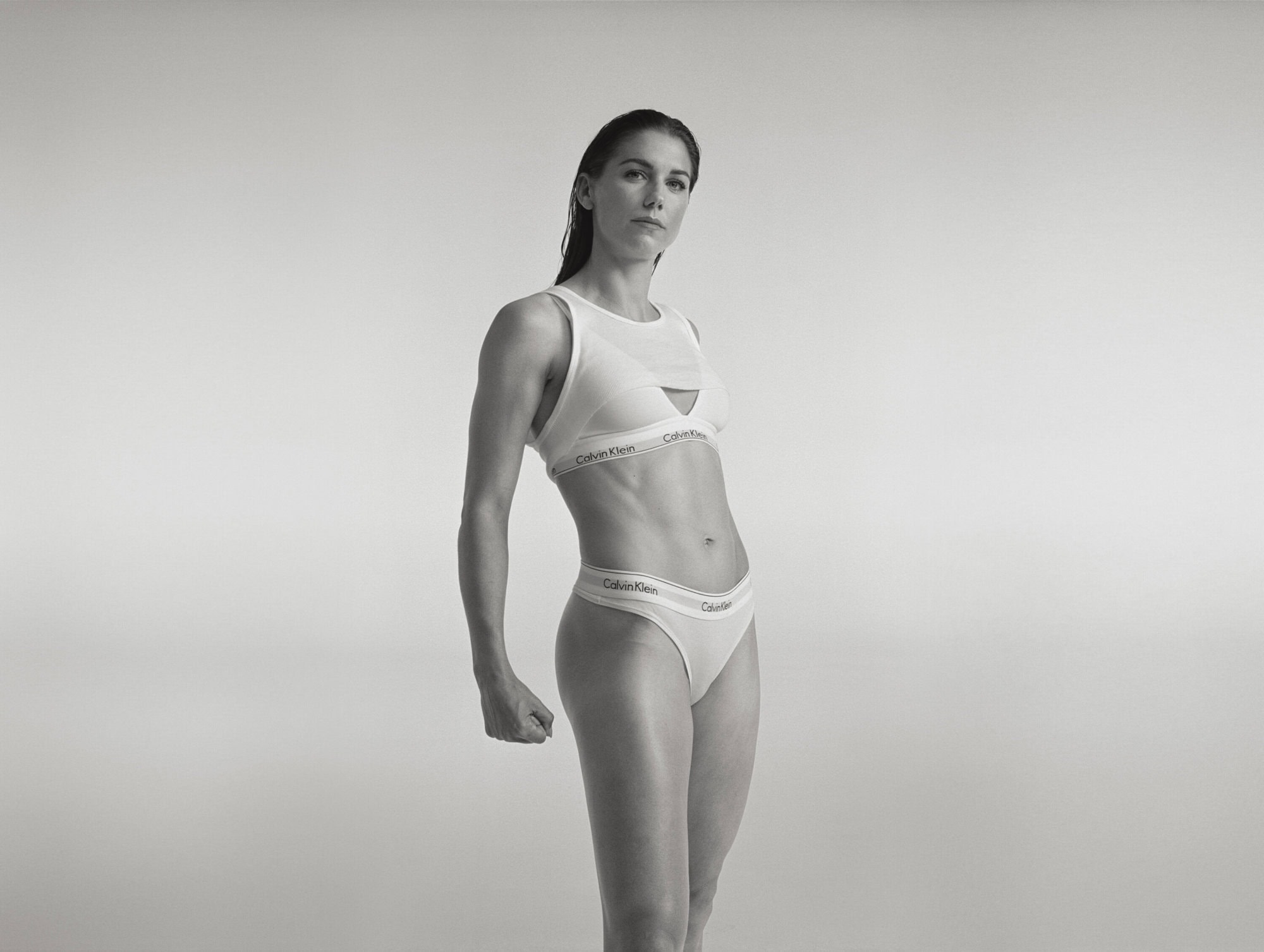 Monochrome image of a muscular woman in Calvin Klein undergarments, posing in front of a bright, neutral background. Fitness or fashion editorial.