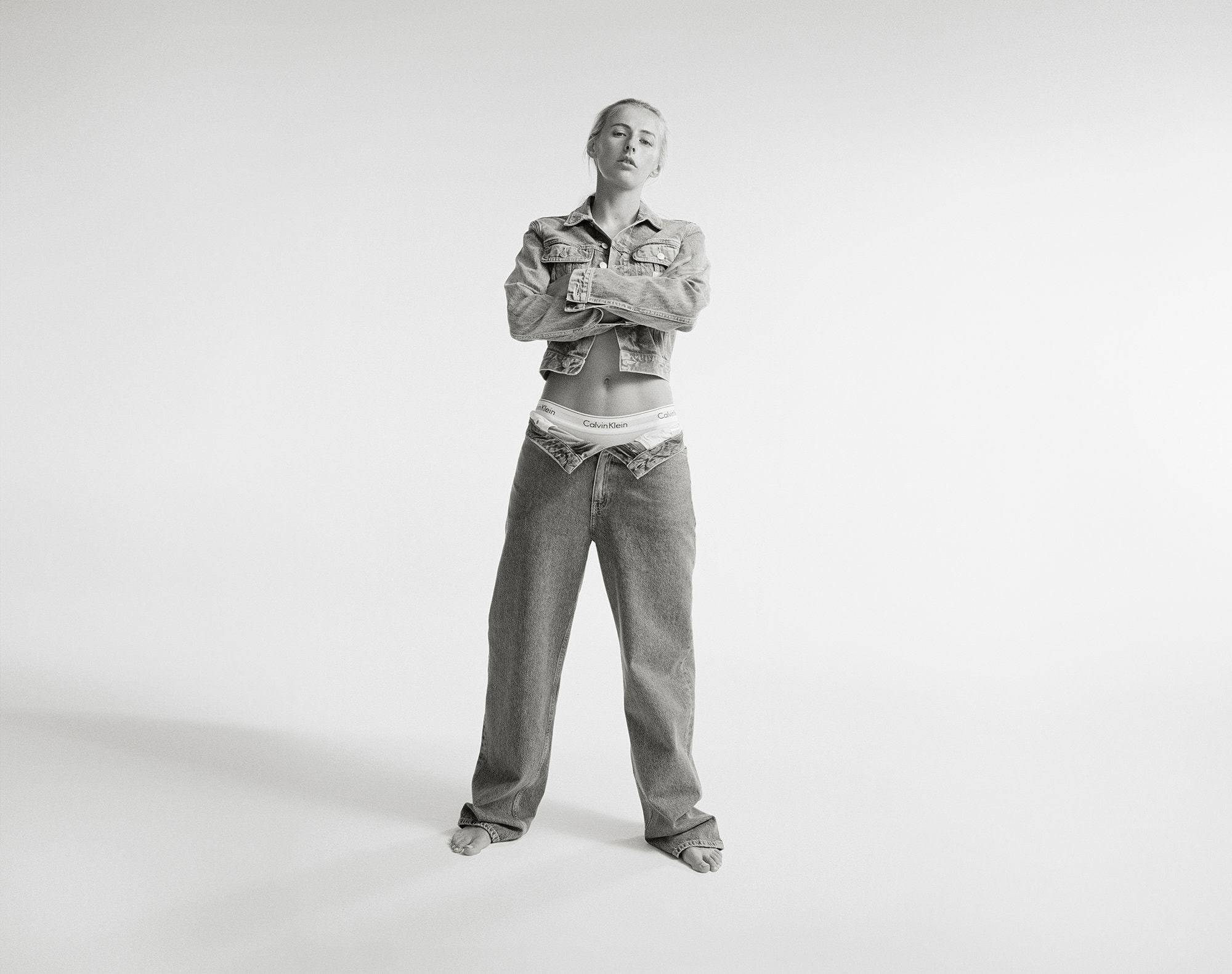 Black and white portrait of a woman in denim jacket and jeans standing against a white backdrop with arms crossed.