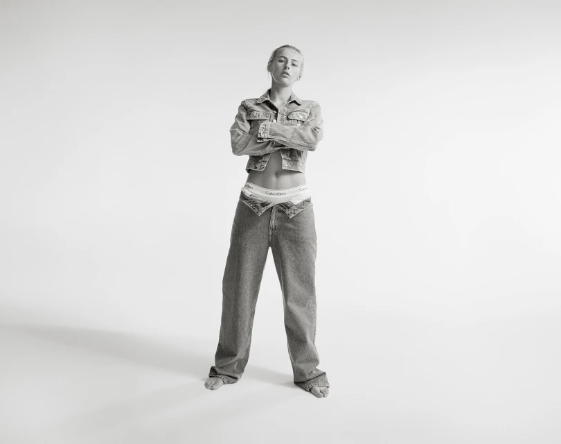 Black and white portrait of a woman in denim jacket and jeans standing against a white backdrop with arms crossed.