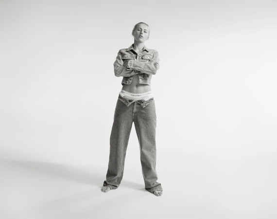 Black and white portrait of a woman in denim jacket and jeans standing against a white backdrop with arms crossed.