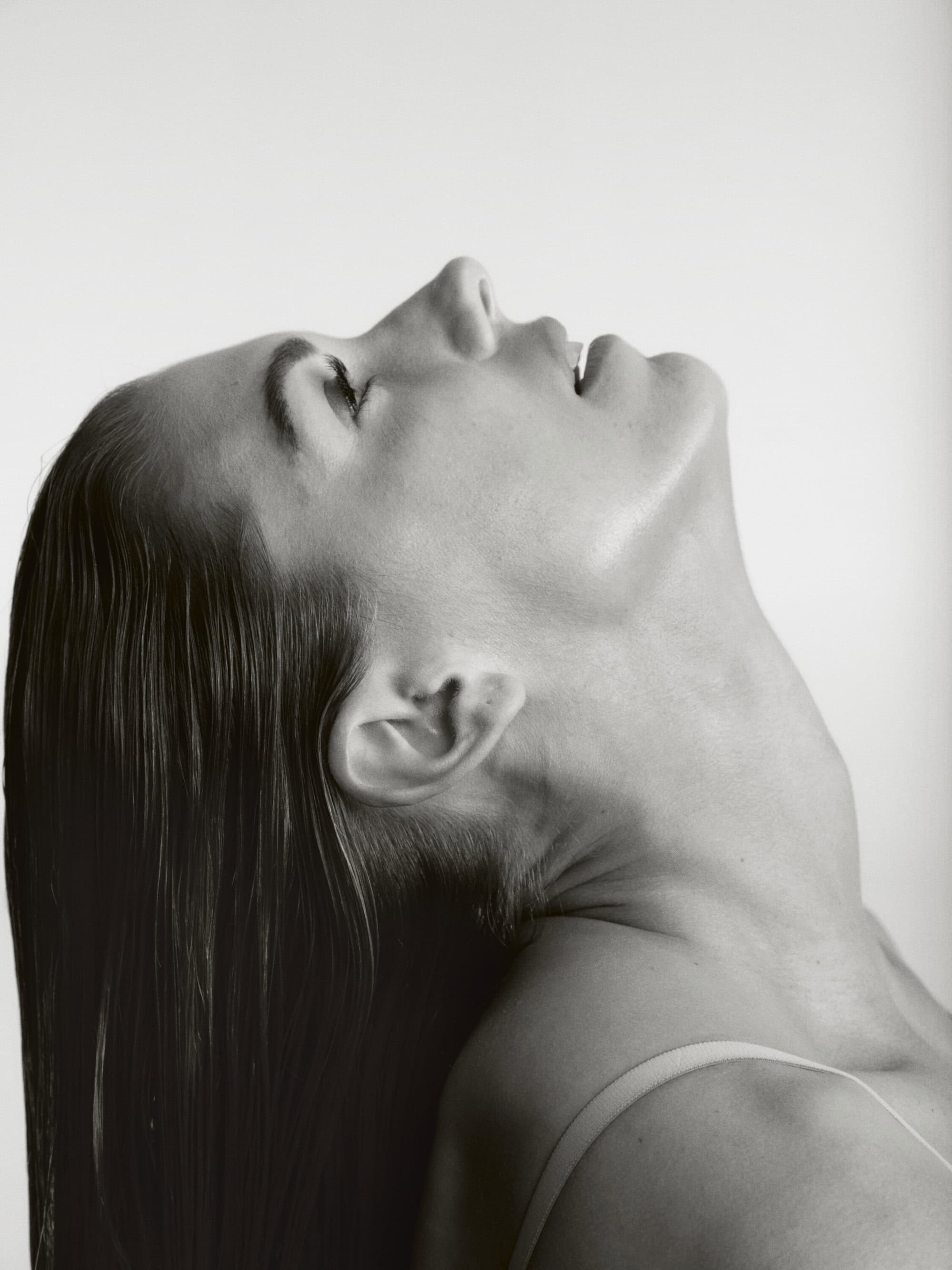 Monochrome portrait of a woman with slicked-back hair looking upwards in a studio setting against a white backdrop.