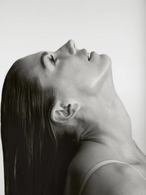 Monochrome portrait of a woman with slicked-back hair looking upwards in a studio setting against a white backdrop.
