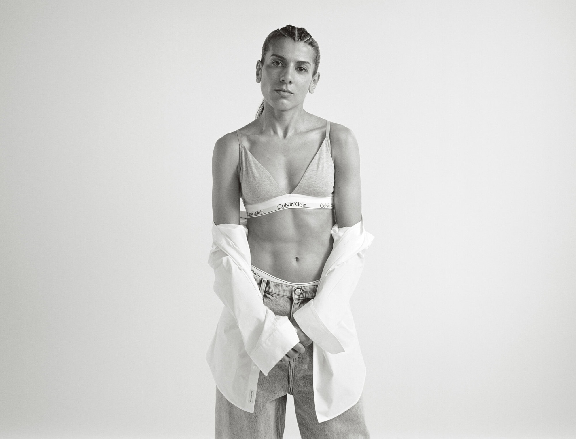Monochrome portrait of a woman in a Calvin Klein bra, unbuttoned shirt, and jeans, posed in a white studio with a serious expression.