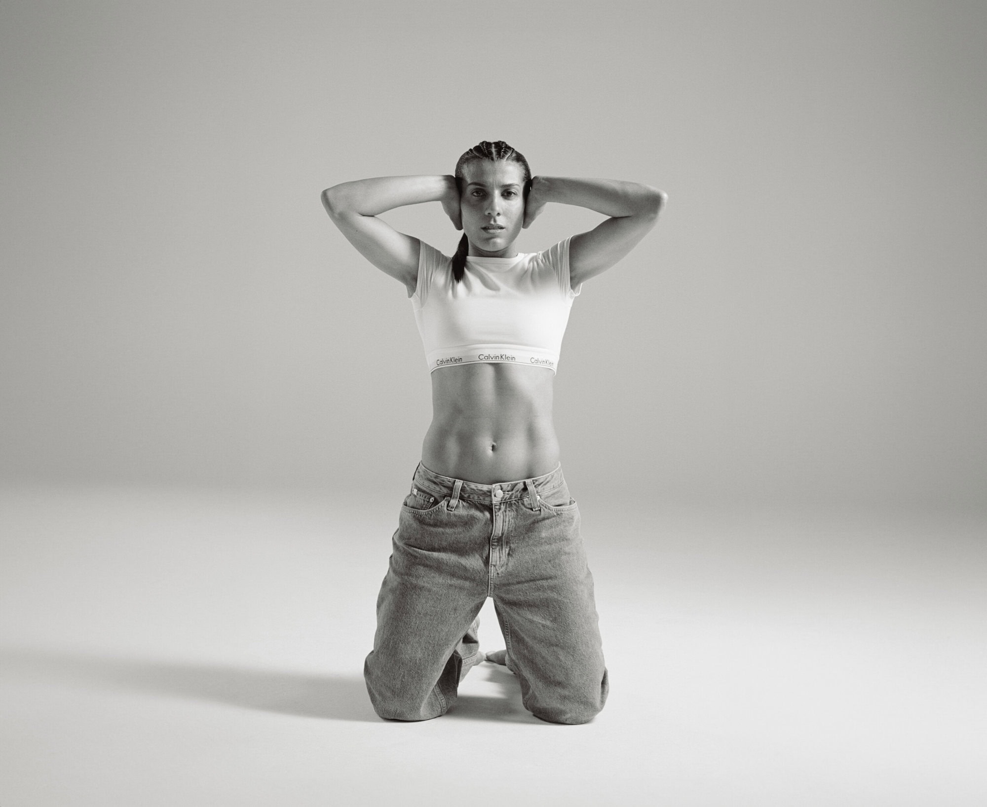 A woman with braided hair kneels in a studio, wearing a white crop top with 'Calvin Klein' on the band and denim jeans, hands behind her head.