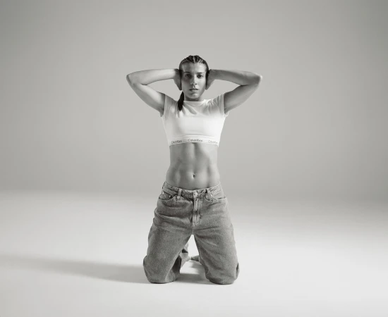 A woman with braided hair kneels in a studio, wearing a white crop top with 'Calvin Klein' on the band and denim jeans, hands behind her head.