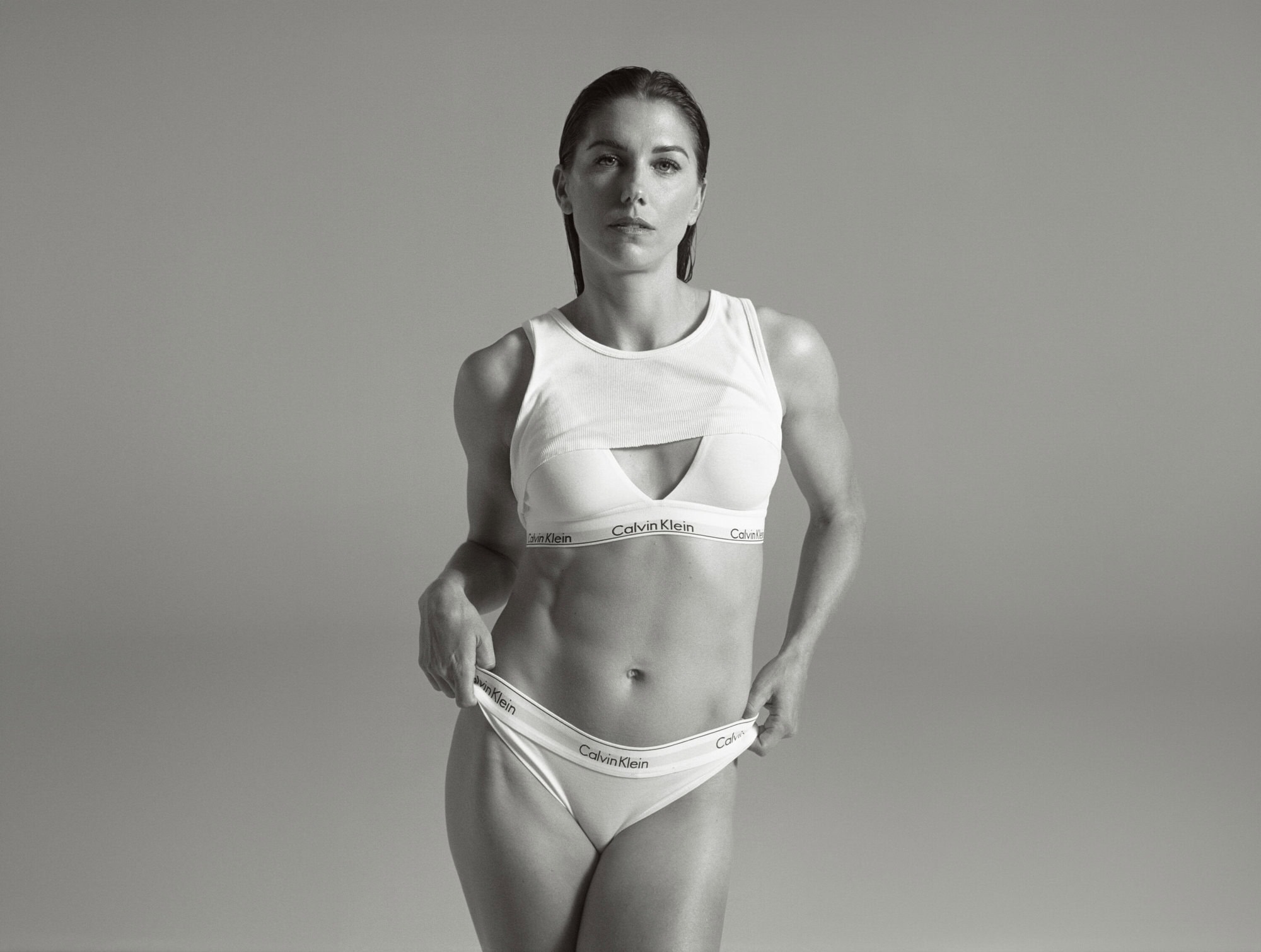 Athletic woman in underwear and sports bra poses in front of grey studio background. Calvin Klein logo is visibly printed on clothing.