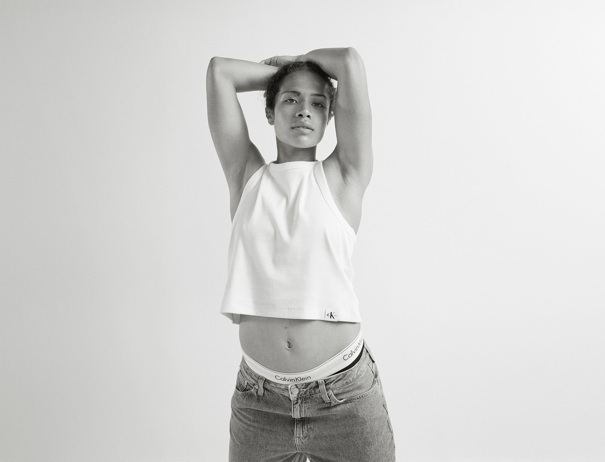 Monochrome shot of a slender adult female in a white tank top and jeans, arms overhead, showcasing Calvin Klein branded underwear.