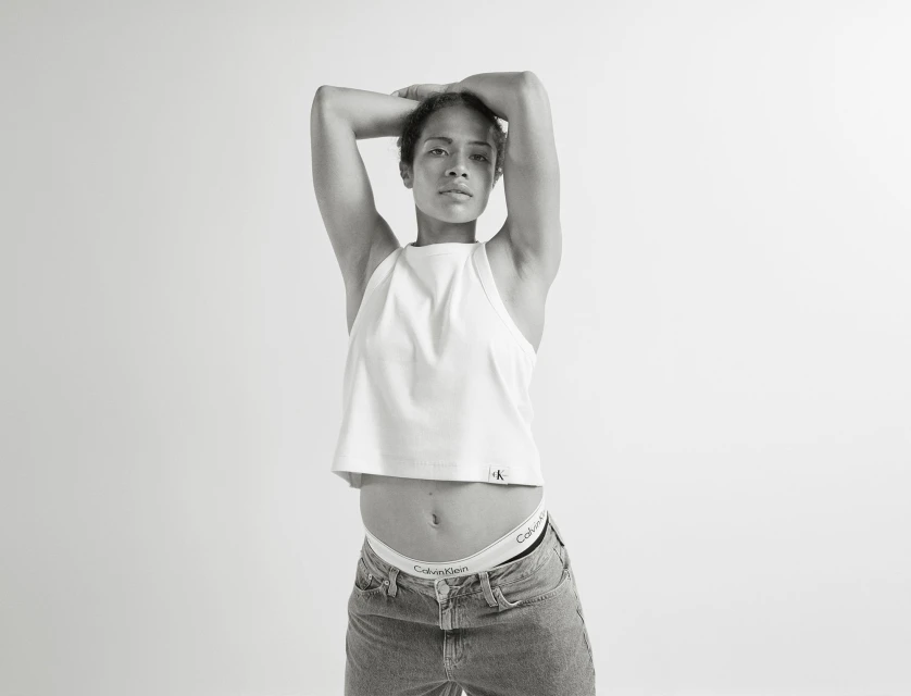 Monochrome shot of a slender adult female in a white tank top and jeans, arms overhead, showcasing Calvin Klein branded underwear.
