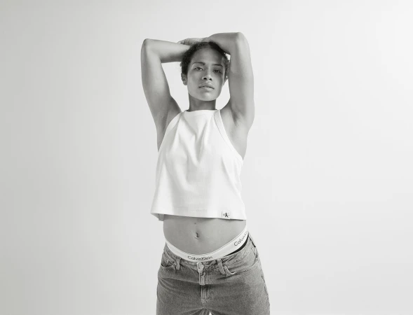 Monochrome shot of a slender adult female in a white tank top and jeans, arms overhead, showcasing Calvin Klein branded underwear.