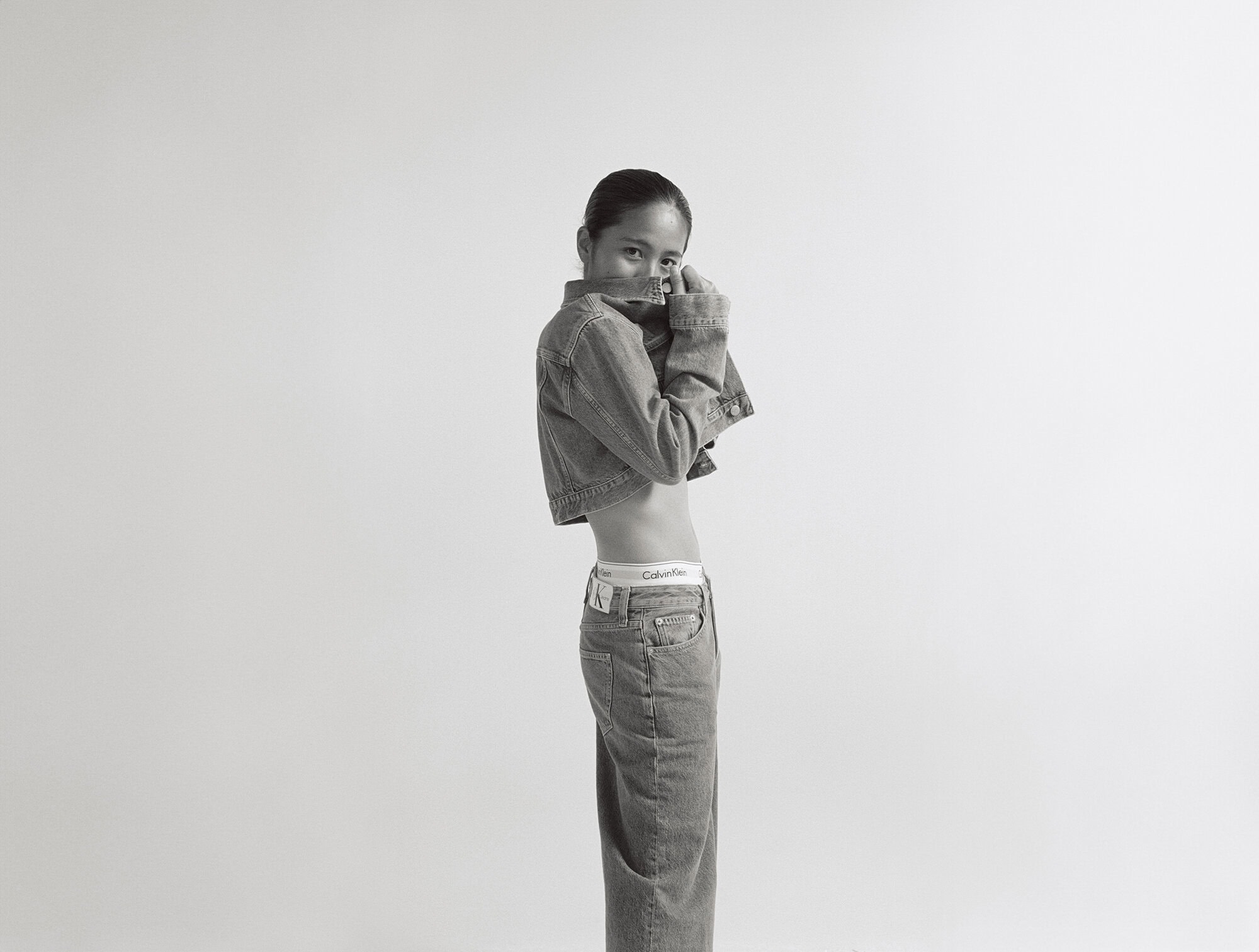 Black-and-white fashion shot of a young woman wearing a denim jacket and jeans, posing in a photo studio against a plain white backdrop.