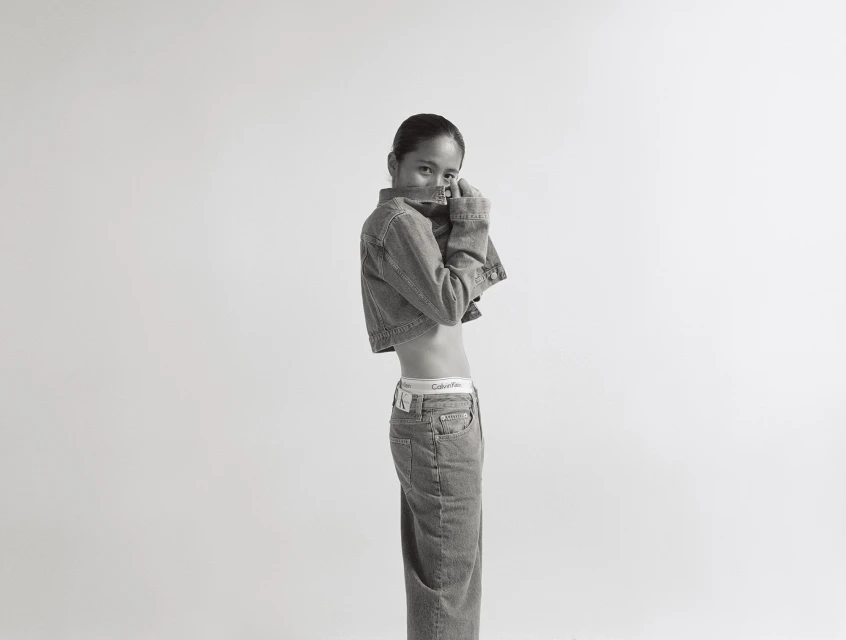 Black-and-white fashion shot of a young woman wearing a denim jacket and jeans, posing in a photo studio against a plain white backdrop.