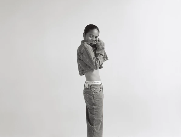 Black-and-white fashion shot of a young woman wearing a denim jacket and jeans, posing in a photo studio against a plain white backdrop.