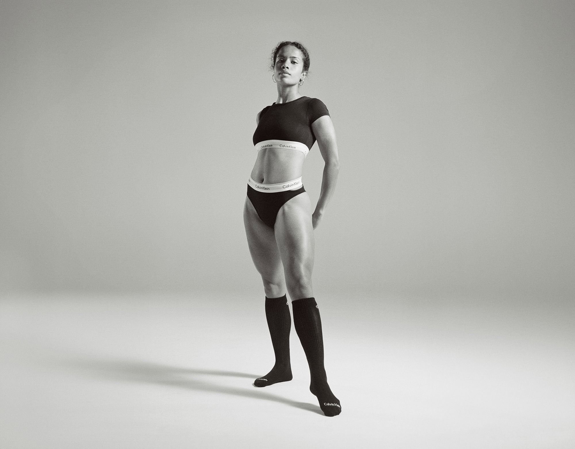 Athletic woman in black crop top and underwear with knee socks stands in a photo studio with grey backdrop; athletic, fit, model.
