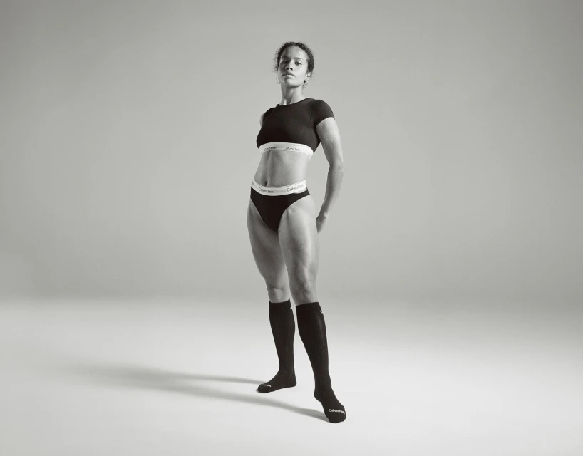 Athletic woman in black crop top and underwear with knee socks stands in a photo studio with grey backdrop; athletic, fit, model.