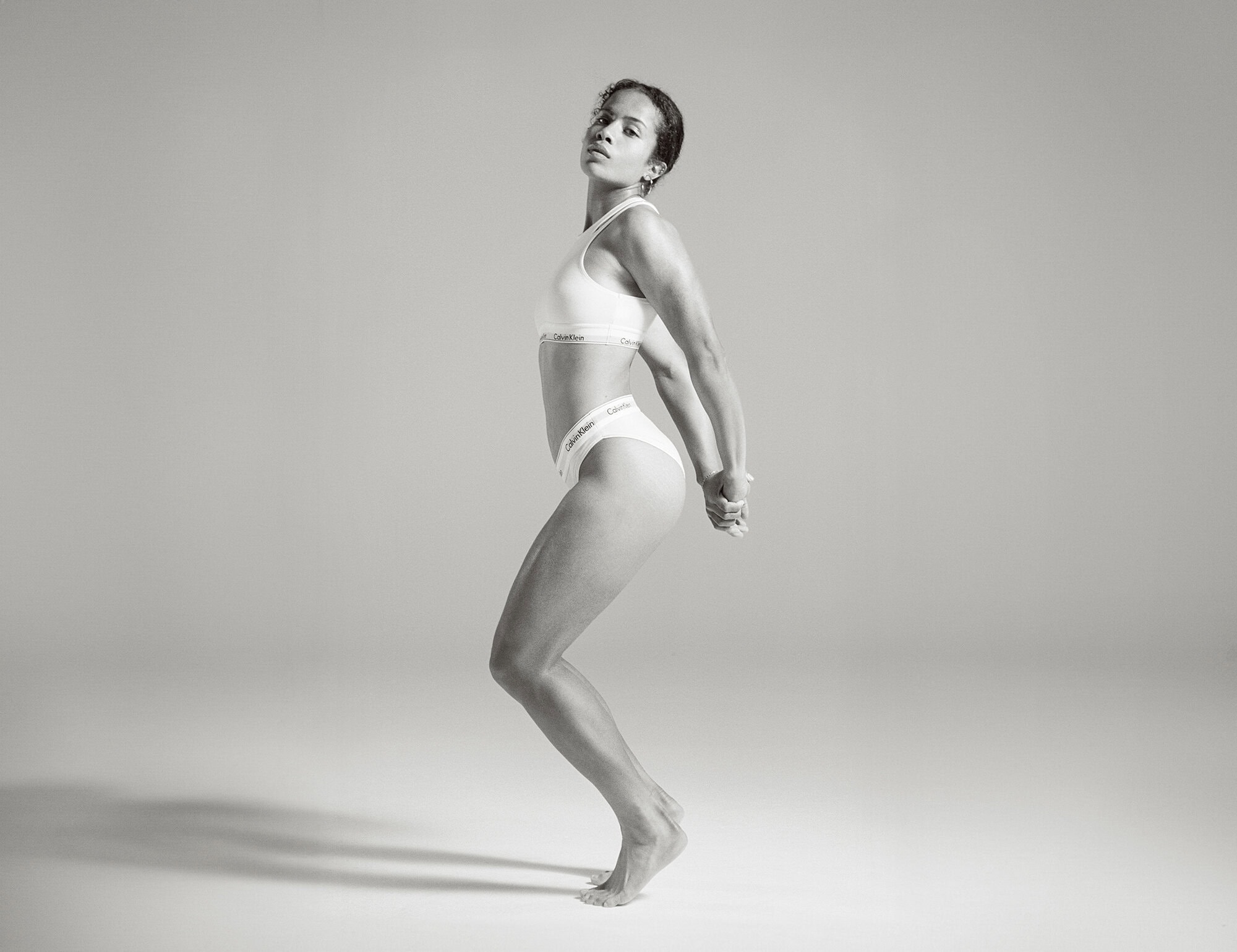 Monochrome studio shot of athletic adult woman in Calvin Klein underwear, posing with hands clasped behind her back in a dynamic stance.