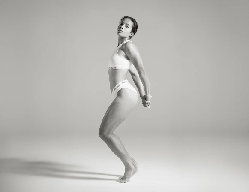 Monochrome studio shot of athletic adult woman in Calvin Klein underwear, posing with hands clasped behind her back in a dynamic stance.